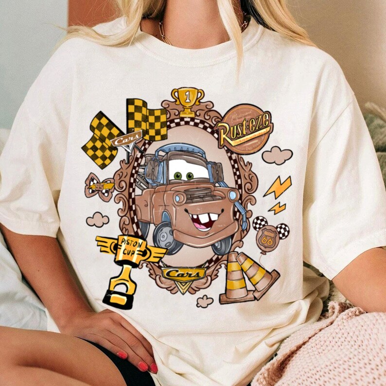 May include: Cream-colored t-shirt featuring a cartoon image of Tow Mater from the movie Cars. The design includes a brown car, checkered flags, lightning bolts, and the words "Rusteze" and "Piston Cup".
