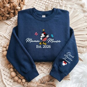 K&ouml;nnte beinhalten: Marineblaues Sweatshirt mit gesticktem Design. Mit "Mama Mouse" Text, Minnie Mouse Grafik, Schmetterlingen und "Est. 2026". Der &Auml;rmel hat Namen und ein Minnie Mouse Symbol.