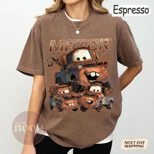 May include: Brown t-shirt with a graphic of the character Mater from the film Cars. The design includes the word "MATER" above the character and three smaller Mater images below. The Cozy Nook logo is visible.