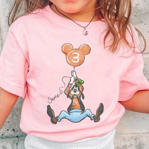 May include: A light pink t-shirt featuring a cartoon of Goofy holding a balloon with the number 3. The balloon is brown and shaped like Mickey Mouse ears. The name "James" is written on the shirt.
