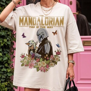 May include: Cream-colored t-shirt featuring a graphic of The Mandalorian and Grogu surrounded by flowers and butterflies. The text "THE MANDALORIAN" and "THIS IS THE WAY" are printed above the characters in gold.