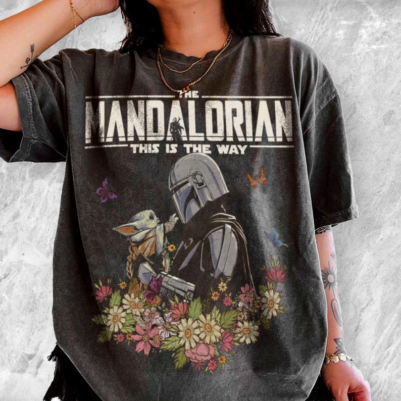 May include: Dark gray t-shirt featuring a graphic of The Mandalorian and Grogu surrounded by flowers and butterflies. The text "THE MANDALORIAN" and "THIS IS THE WAY" are printed across the top of the design.
