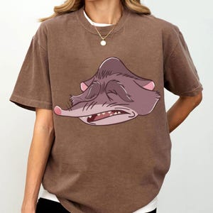 Peut inclure: T-shirt marron avec une illustration de dessin anim&eacute; d'un rongeur grincheux aux yeux ferm&eacute;s et &agrave; la bouche l&eacute;g&egrave;rement ouverte. Le graphisme est dans les tons de violet, rose et blanc. Le t-shirt a des manches courtes et un col rond.