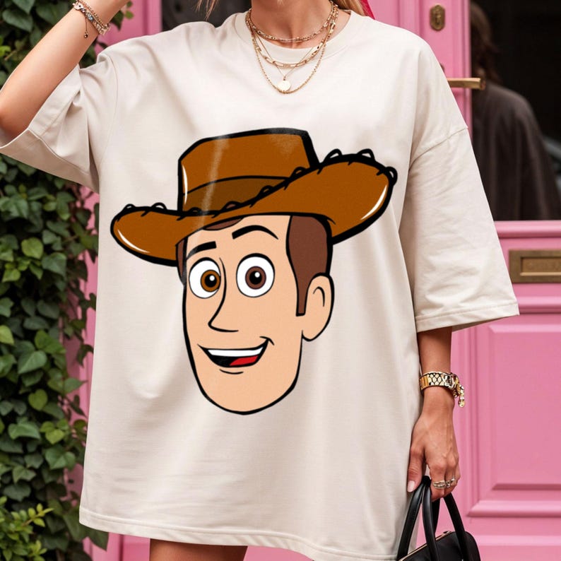 May include: Cream-colored t-shirt featuring a cartoon illustration of a character wearing a brown cowboy hat. The character has brown hair, brown eyes, and a smiling expression. The design is centered on the front of the shirt.