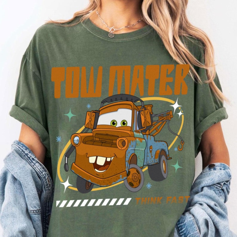 May include: An olive green t-shirt featuring a graphic of the character Tow Mater from the movie Cars. The design includes the text TOW MATER in orange, the character in brown and blue, and the text THINK FAST in orange with white stripes.