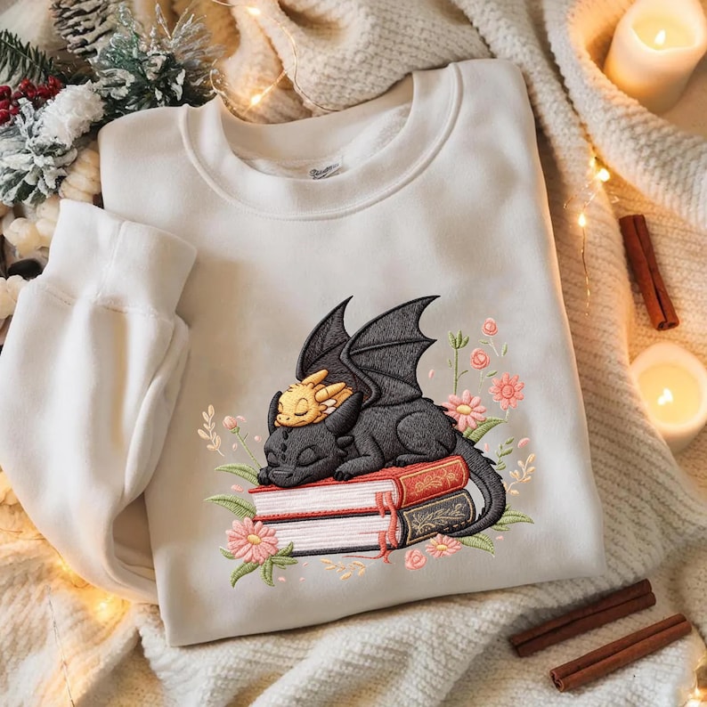 May include: White sweatshirt featuring an embroidered design of a black dragon and a yellow dragon resting on a stack of books, surrounded by pink flowers and green leaves. The sweatshirt has a crew neck and long sleeves.