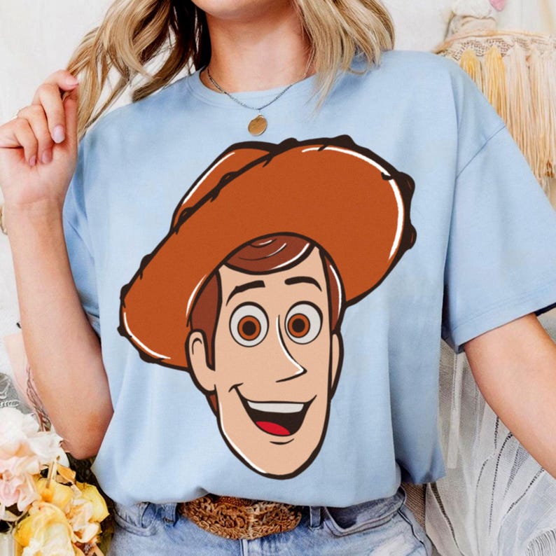 May include: Light blue t-shirt featuring a cartoon illustration of Woody from Toy Story. The graphic shows Woody's face with a brown cowboy hat, brown hair, and a smiling expression. The shirt is a casual wear item.