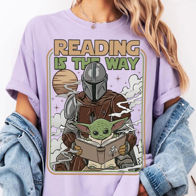 Puede incluir: Una camiseta lila claro con un gr&aacute;fico de The Mandalorian y Grogu leyendo un libro. El texto "READING IS THE WAY" se muestra sobre los personajes en un estilo retro. El dise&ntilde;o incluye un sol, estrellas y humo.