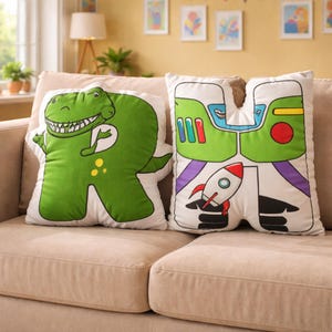 Puede incluir: Dos cojines decorativos sobre un sof&aacute; beige. Un coj&iacute;n tiene forma de letra "R" y presenta un dinosaurio verde. El otro coj&iacute;n tiene forma de letra "H" y tiene un cohete y detalles en verde y rojo.
