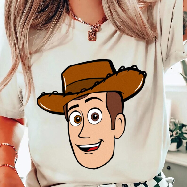 May include: A cream-colored t-shirt featuring a cartoon illustration of a smiling cowboy character with a brown cowboy hat. The character has brown hair, a tan face, and a friendly expression.