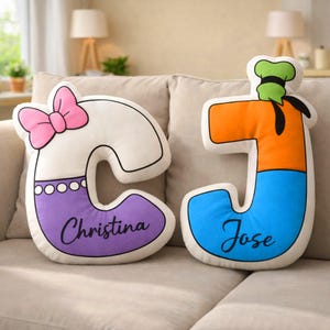 Op de afbeelding: Twee kussens in de vorm van letters. De letter "C" is wit met een roze strik, een paarse basis met de naam "Christina". De letter "J" is oranje en blauw met de naam "Jose" en een groene hoed.