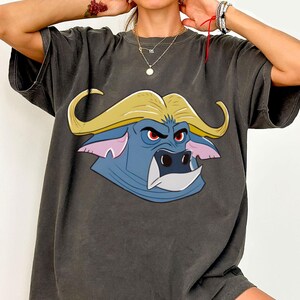 May include: Dark gray t-shirt featuring a cartoon buffalo head with yellow hair, blue face, pink ears, and red eyes. The buffalo has a white tooth and a stern expression. The shirt is a casual wear item.