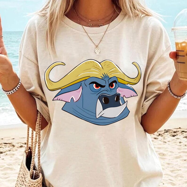 May include: A beige t-shirt featuring a cartoon buffalo head with yellow horns, blue face, and red eyes. The buffalo graphic is centered on the shirt. The shirt is a casual style, suitable for everyday wear.