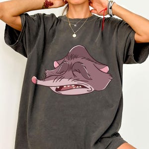 Peut inclure: T-shirt gris fonc&eacute; avec une illustration de dessin anim&eacute; d'un visage d'animal endormi, violet-gris, avec des accents roses. Le dessin est centr&eacute; sur le devant. Le t-shirt a des manches courtes et une coupe d&eacute;contract&eacute;e.