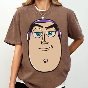 May include: Brown t-shirt featuring a cartoon illustration of a character's face with blue eyes, purple accents, and a smiling expression. The shirt has short sleeves and a crew neck.