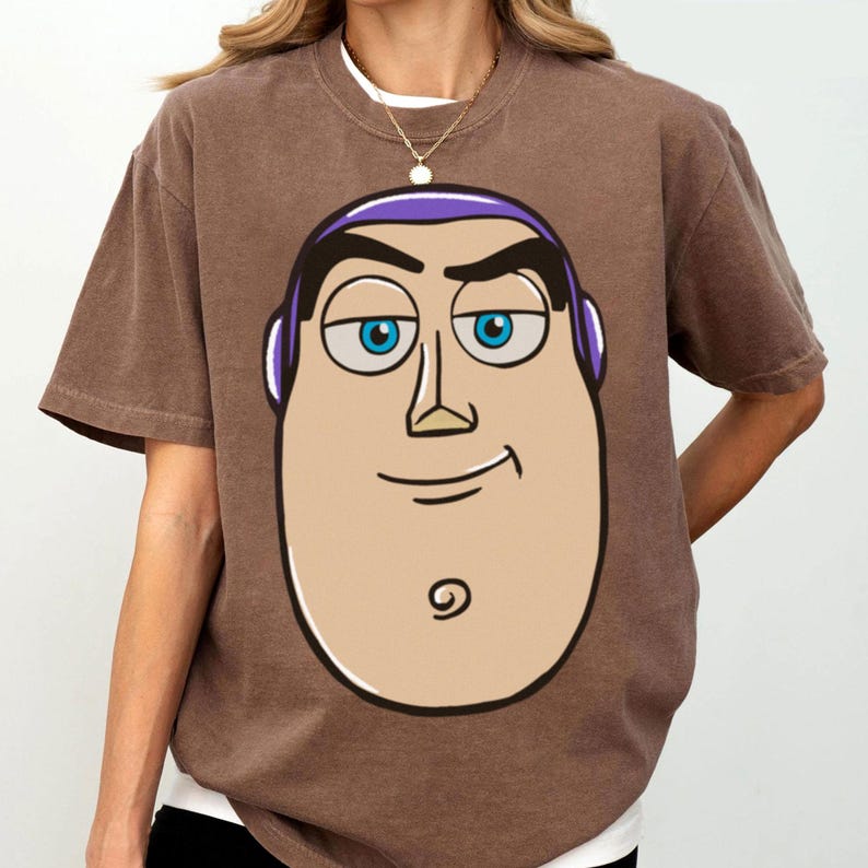 May include: Brown t-shirt featuring a cartoon illustration of a character's face with blue eyes, purple accents, and a smiling expression. The shirt has short sleeves and a crew neck.