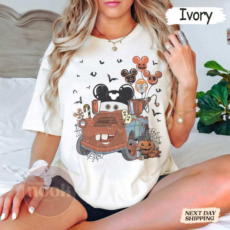 May include: Ivory-colored t-shirt featuring a Halloween-themed graphic. The design includes a cartoon truck with Mickey Mouse ears, balloons, pumpkins, ghosts, and bats. The text "Ivory" is printed on the top right.