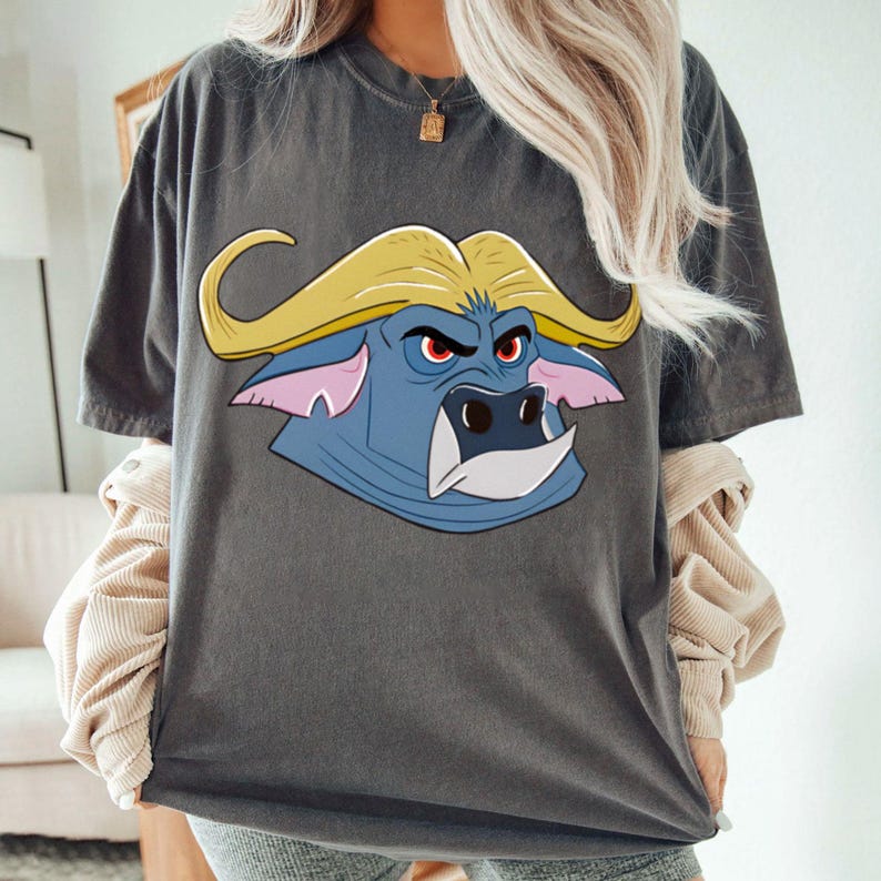 May include: Dark gray t-shirt featuring a cartoon buffalo head with yellow horns, blue face, and red eyes. The buffalo graphic is centered on the front of the shirt. The shirt has short sleeves and a crew neck.