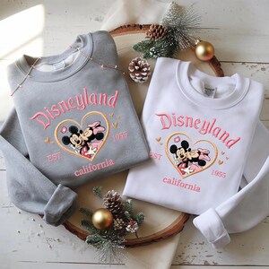 Peut inclure: Deux sweatshirts, un gris et un blanc, avec le logo Disneyland brod&eacute; en rose. Le motif comprend Mickey et Minnie Mouse dans un c&oelig;ur, avec "EST 1955 California" en dessous. Pr&eacute;sent&eacute;s sur une planche en bois avec des d&eacute;corations festives.