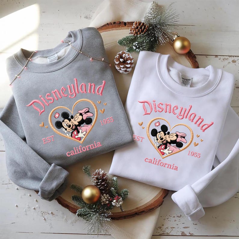 Peut inclure: Deux sweatshirts, un gris et un blanc, avec le logo Disneyland brod&eacute; en rose. Le motif comprend Mickey et Minnie Mouse dans un c&oelig;ur, avec "EST 1955 California" en dessous. Pr&eacute;sent&eacute;s sur une planche en bois avec des d&eacute;corations festives.