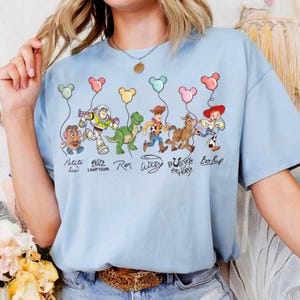 May include: Pale blue t-shirt with a vibrant graphic of Toy Story characters, each holding Mickey Mouse-shaped balloons. The design includes Mr. Potato Head, Buzz Lightyear, Rex, Woody, Bullseye, and Bo Peep, with their names beneath them.