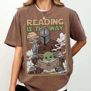 Puede incluir: Camiseta marr&oacute;n con un dise&ntilde;o retro de The Mandalorian y Grogu leyendo un libro, con el texto "READING IS THE WAY". El dise&ntilde;o incluye un atardecer y nubes, creando una est&eacute;tica vintage.
