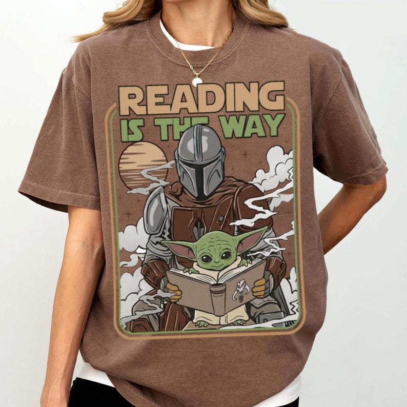 Puede incluir: Camiseta marr&oacute;n con un dise&ntilde;o retro de The Mandalorian y Grogu leyendo un libro, con el texto "READING IS THE WAY". El dise&ntilde;o incluye un atardecer y nubes, creando una est&eacute;tica vintage.