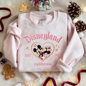 Peut inclure: Un sweat-shirt rose clair avec le mot "Disneyland" brod&eacute; en rose. En dessous, un motif en forme de c&oelig;ur avec Mickey et Minnie Mouse, avec "EST 1955 California" brod&eacute; en dessous. Le sweat-shirt est pli&eacute;.
