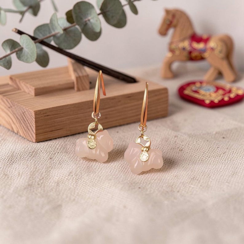 May include: A pair of gold-colored drop earrings with pink, carved, animal-shaped pendants. The earrings are displayed on a textured, off-white surface, with a wooden incense holder and decorative items in the background.