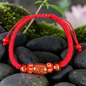 Puede incluir: Una pulsera de cordón trenzado rojo con una cuenta central en forma de barril con la palabra PEACE grabada en oro. La pulsera incluye dos cuentas rojas más pequeñas, una con un símbolo de infinito y otra con una paloma, separadas por nudos dorados.