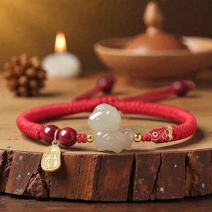 May include: Red braided bracelet with a white jade figurine, red beads, and a gold charm with Chinese characters. The bracelet has a fish-shaped accent and is displayed on a wooden surface.