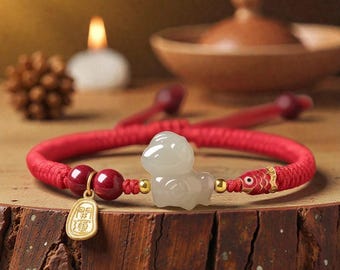 May include: Red braided bracelet featuring a white jade figurine, red beads, and a gold charm with Chinese characters. The bracelet has a fish-shaped accent and is displayed on a wooden surface.