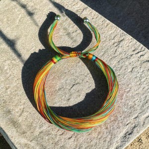 Peut inclure: Un bracelet coloré composé de nombreux fils rouges, oranges, jaunes, verts et bleus. Le bracelet est noué au centre et comporte des perles argentées aux extrémités des liens réglables. Le bracelet est posé sur une surface en pierre.