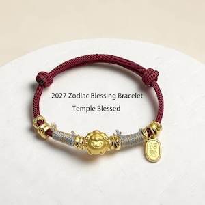 May include: A maroon cord bracelet featuring a central gold-toned sheep charm, flanked by light blue and gold thread-wrapped sections, gold-toned beads, and a small rectangular gold-toned tag with Chinese characters. Text reads: 2027 Zodiac Blessing Bracelet Temple Blessed.