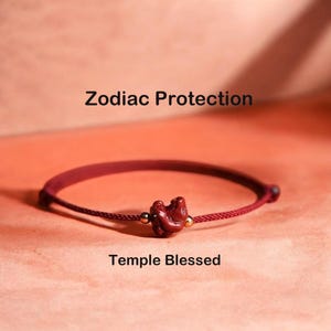 Könnte beinhalten: Ein dunkelrotes Kordelarmband mit einem zentralen, geschnitzten roten Tieranhänger, der von zwei kleinen goldfarbenen Perlen flankiert wird. Der Text Zodiac Protection und Temple Blessed ist über und unter dem geflochtenen Armband zu sehen.