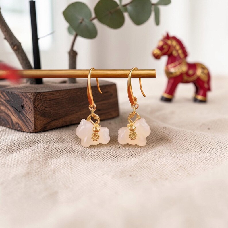 May include: A pair of gold-colored earrings with a light pink, horse-shaped pendant. The earrings are displayed on a gold bar with a wooden base. A red horse figurine is in the background.