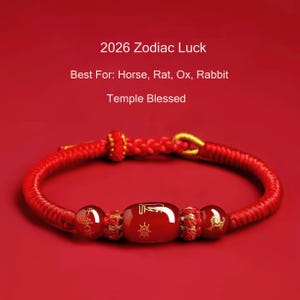 Puede incluir: Pulsera trenzada roja con tres cuentas decorativas y una cuenta rectangular central. La pulsera tiene detalles dorados y está sobre un fondo rojo. El texto de la imagen dice "2026 Zodiac Luck" y "Temple Blessed".