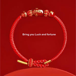 May include: A red braided bracelet with gold accents and decorative beads. The beads have gold detailing and Chinese characters. The text "Bring you Luck and fortune" is displayed on a red background.