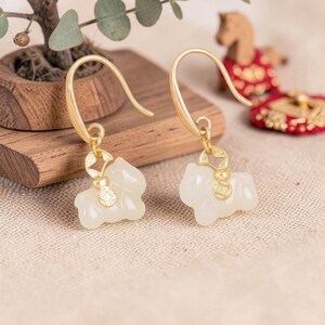 May include: A pair of gold-colored hook earrings. Each earring features a small, off-white, horse-shaped charm with a gold-colored coin detail. The earrings are displayed on a neutral-toned surface.