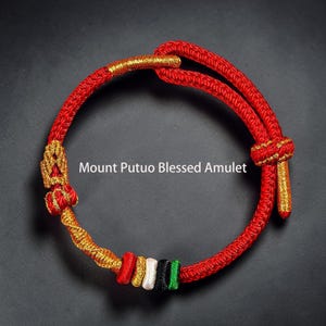Puede incluir: Una pulsera de cordón rojo con detalles de hilo dorado, un cierre de nudo corredizo y pequeñas cuentas cilíndricas en rojo, blanco, negro y verde. El texto Mount Putuo Blessed Amulet está impreso en blanco junto a la estructura circular.