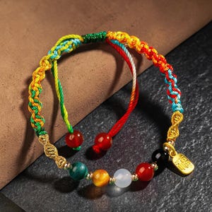 Peut inclure: Un bracelet tressé multicolore avec des perles de pierres précieuses. Le bracelet présente un arc-en-ciel de couleurs dans le cordon tressé et comprend des perles rouges, vertes, jaunes et blanches. Un petit charme doré est suspendu au bracelet.