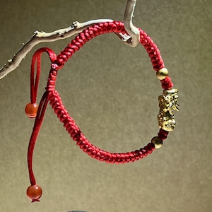 Op de afbeelding: Rode gevlochten armband met gouden accenten en een gouden bedel. De armband is verstelbaar met een schuifknoop en heeft twee kleine oranje kralen. De gouden bedel is een gedetailleerde figuur.