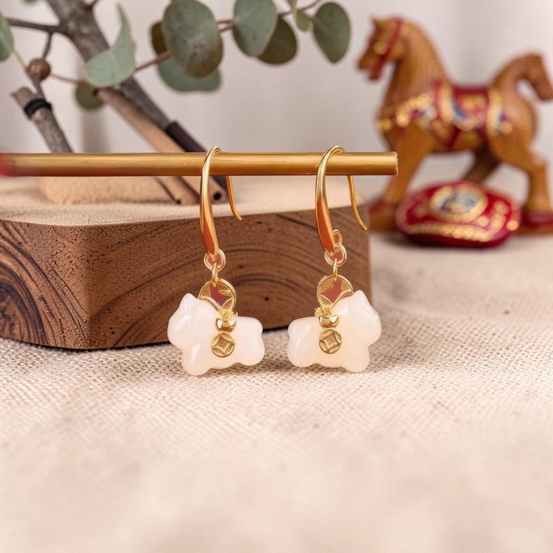 May include: Gold-tone earrings featuring a horse-shaped pendant in a light pink hue. The earrings have a hook closure and a small, round charm with a gold-colored design. The earrings are displayed on a wooden surface.