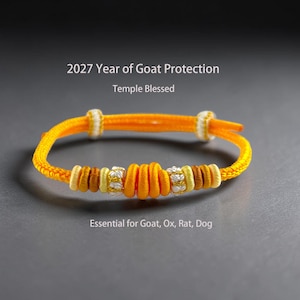 May include: An orange cord bracelet featuring a central knot, flanked by smaller yellow, brown, and white textured beads. The text reads: 2027 Year of Goat Protection, Temple Blessed, Essential for Goat, Ox, Rat, Dog. The bracelet has two sliding knots.