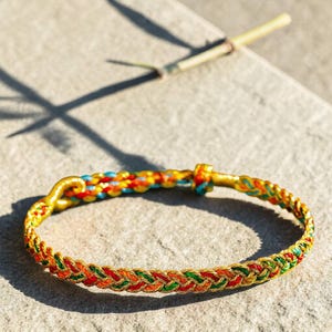 May include: A braided bracelet with a gold clasp and accents. The bracelet features a woven pattern of red, green, and orange threads, with sections of blue and yellow. A bamboo stalk lies in the background.