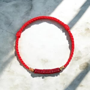 Puede incluir: Una pulsera de cordón trenzado de color rojo brillante que presenta una serie de cinco cuentas rectangulares rojas con caracteres en relieve en el centro, flanqueadas por dos cuentas más pequeñas con patrones multicolores a cada lado.