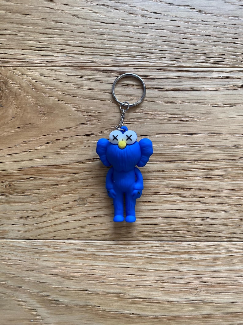 May include: A blue keychain featuring a cartoon character with X-shaped eyes and a yellow nose. The figure has large, rounded ears and is attached to a silver keyring. The keychain is set against a wooden background.