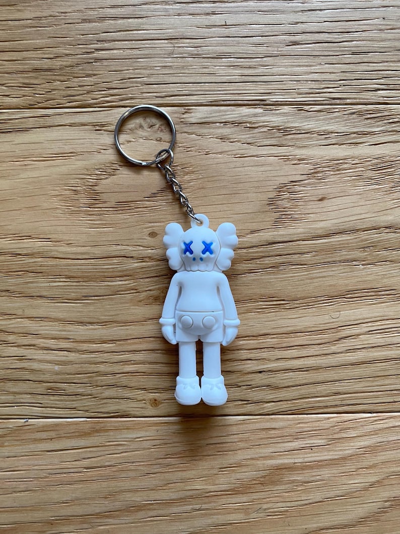May include: A white keychain featuring a stylized figure with blue 'XX' eyes. The figure has a rounded head, small ears, and a button-like detail on the chest. The keychain includes a silver-colored ring and chain, and is set against a wooden background.
