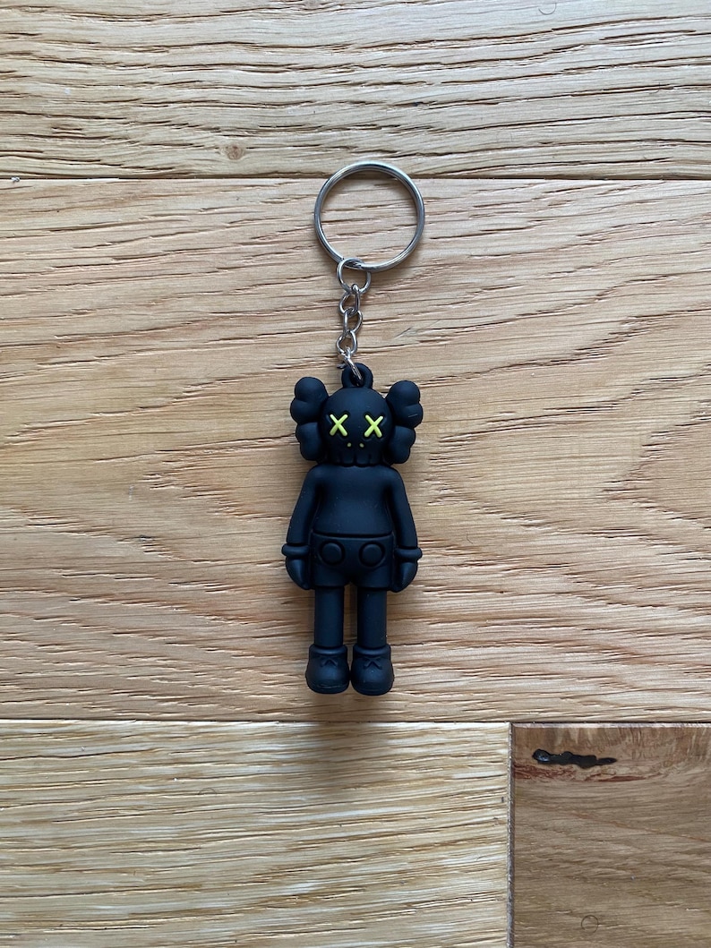 Pode incluir: Chaveiro preto com uma figura estilizada com olhos riscados e cabe&ccedil;a redonda. A figura tem corpo, cal&ccedil;&otilde;es e sapatos pretos, com olhos verde brilhante. O chaveiro inclui um anel e uma corrente prateados.