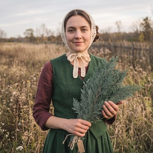 May include: A dark green vest worn over a maroon long-sleeved shirt, paired with a cream-colored crocheted bonnet tied under the chin. The garment is held alongside a bundle of green cedar branches, featuring a small brown rectangular tag attached.